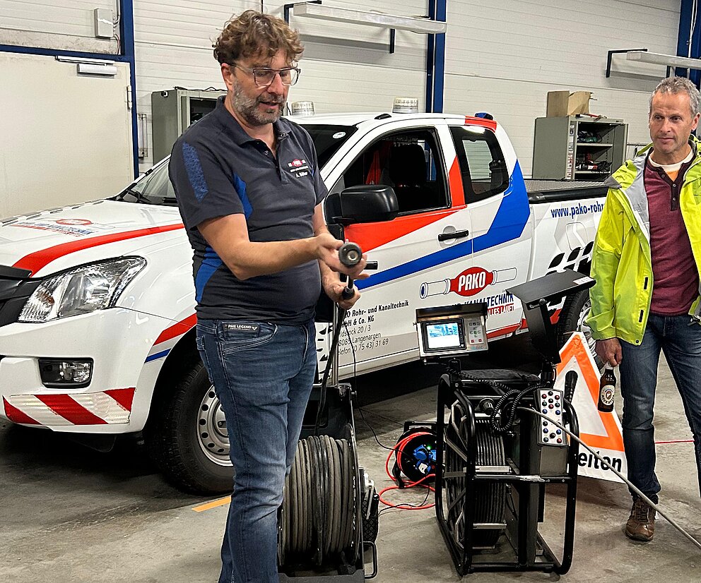 Betriebsbesichtigung bei PAKO Alexander Böhm von PAKO präsentiert ein Kanalreinigugnsgerät, ein Mann in gelber Jacke schaut zu. Im Hintergrund befindet sich ein Dienstfahrzeug.