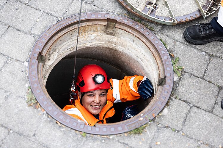 Inspektion im Kanalschacht PAKO-Mitarbeiter mit Helm und Schutzkleidung schaut aus einem geöffneten Kanalschacht, umgeben von Pflastersteinen.