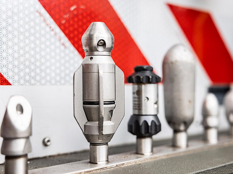 Aufsätze für Kanalreinigung Verschiedene Reinigungsdüsen für die Kanalreinigung aufgereiht in Halterungen, im Hintergrund eine rot-weiße Warntafel.