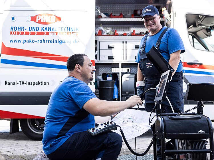 Zwei PAKO-Mitarbeiter in blauer Arbeitskleidung führen Wartungsarbeiten an einem offenen Servicefahrzeug durch, Ausrüstung ist sichtbar.