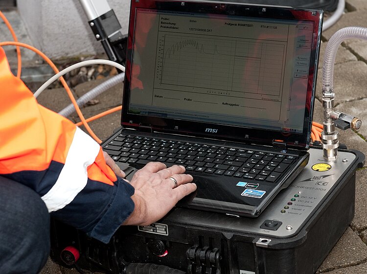 PAKO-Mitarbeiter in Warnweste analysiert Dichtheitsprüfungsdaten am Laptop mit angeschlossenem Messgerät.