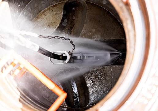 Kanalreinigung durch Schachtdeckel Blick in einen offenen Schacht mit Reinigungsgerät, das im Kanalrohr arbeitet; Details der Rohrreinigung sichtbar.