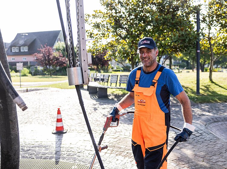 Bauarbeiter bei Schachtarbeit PAKO-Mitarbeiter in oranger Warnkleidung arbeitet mit einem Kollegen an einem geöffneten Schachtdeckel auf einer Straße.